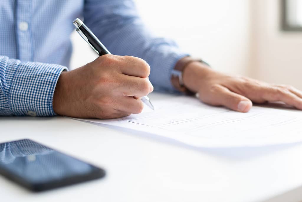 A man, pen in hand, about to sign a document. There is a phone on the table next to him.