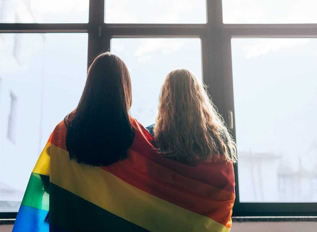 A lesbian couple looking out a window, with a rainbow flag draped over both of their shoulders, thinking about same-sex marriage and inheritance.