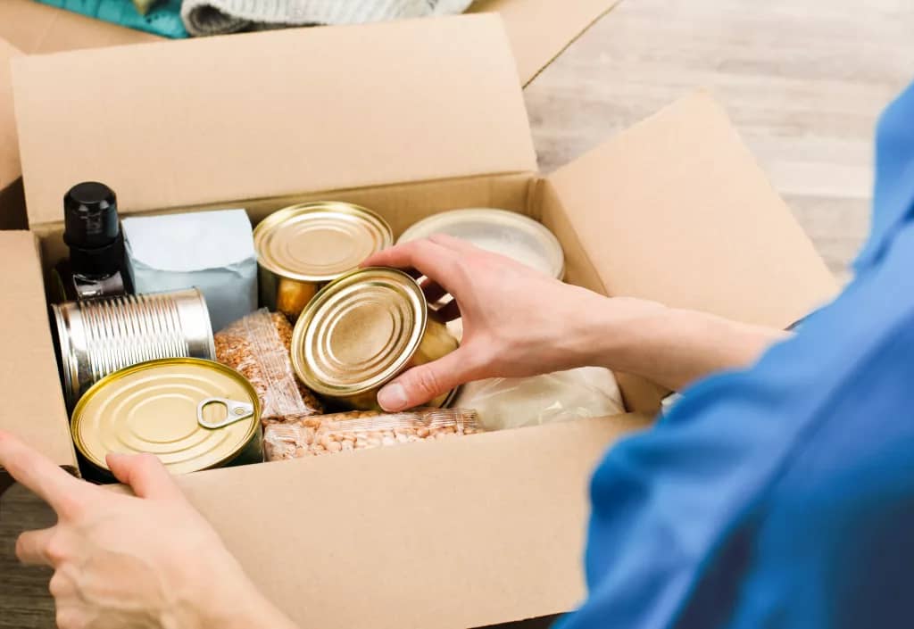 A volunteer filling a box with donations for a food bank - in the box are tins, beans and a bottle.