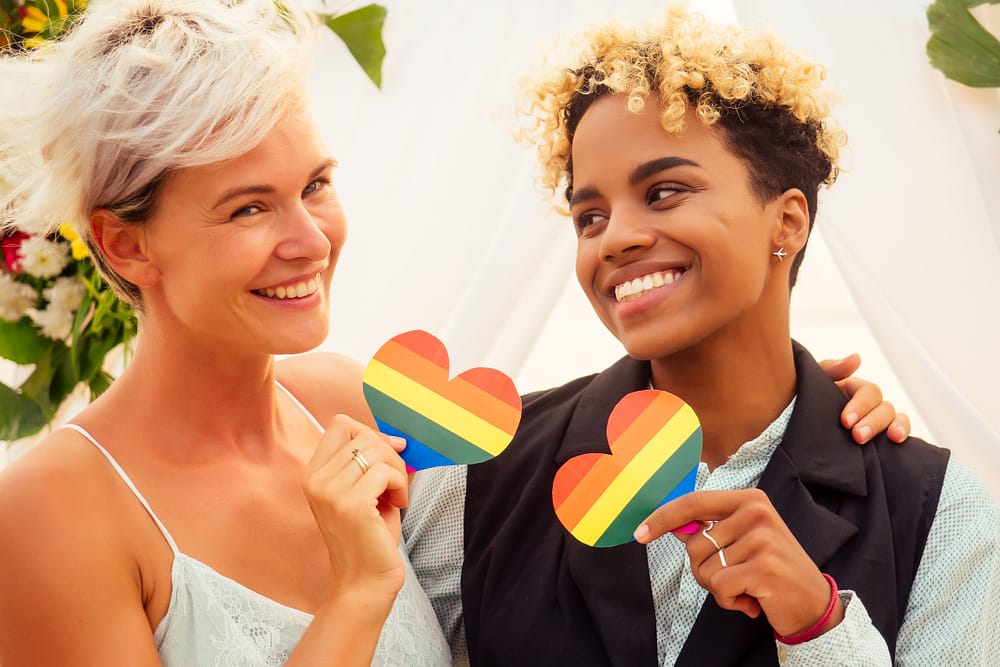 A same-sex couple on their wedding day, holding up little cut-outs of rainbow hearts. One is wearing a white dress, the other a suit jacket.