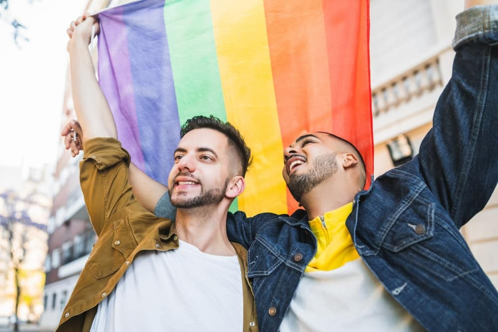 A gay couple, each with an arm around the other's shoulders, holding up a rainbow flag and smiling happily